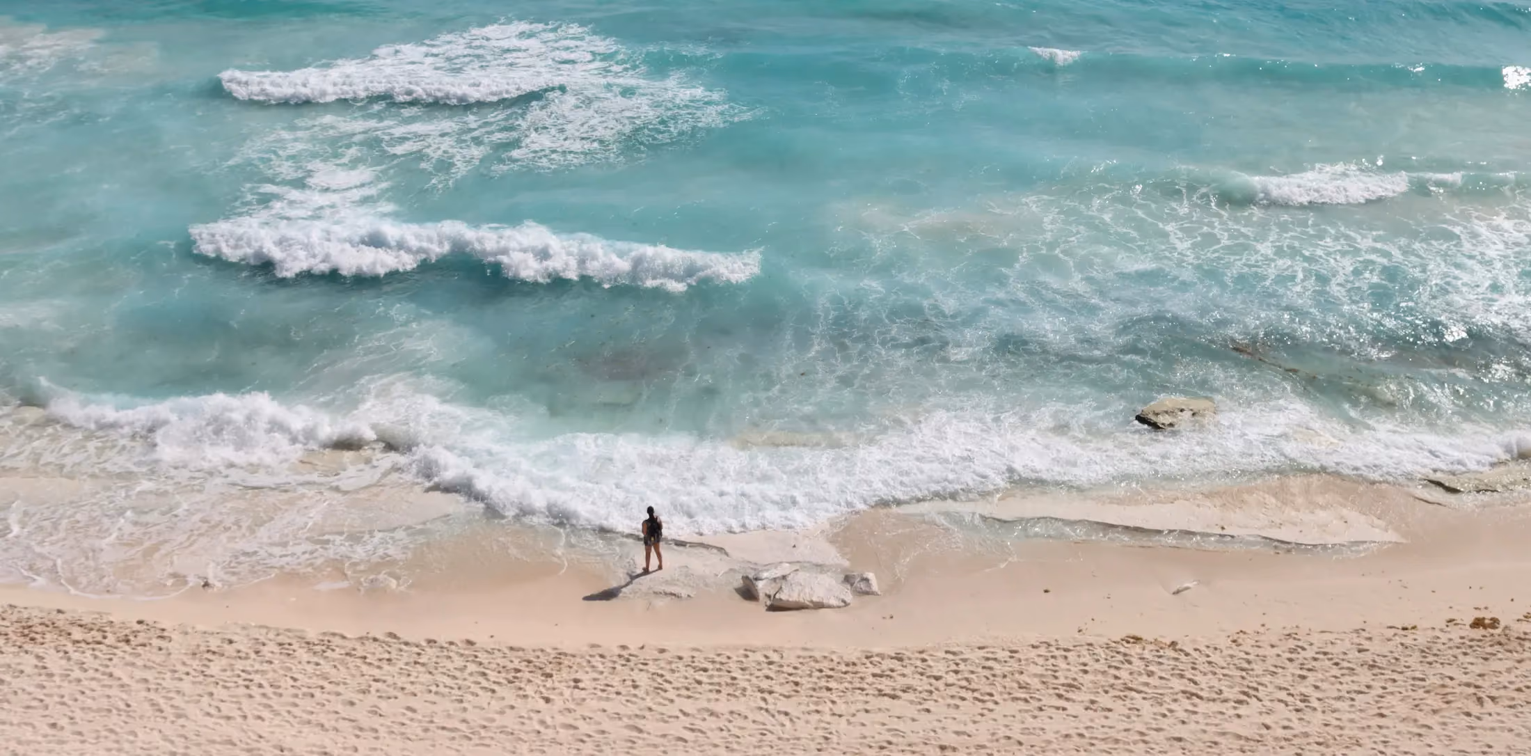 A lone person standing on a sandy beach near gentle turquoise ocean waves with white foam, scattered rocks, and clear shallow water.