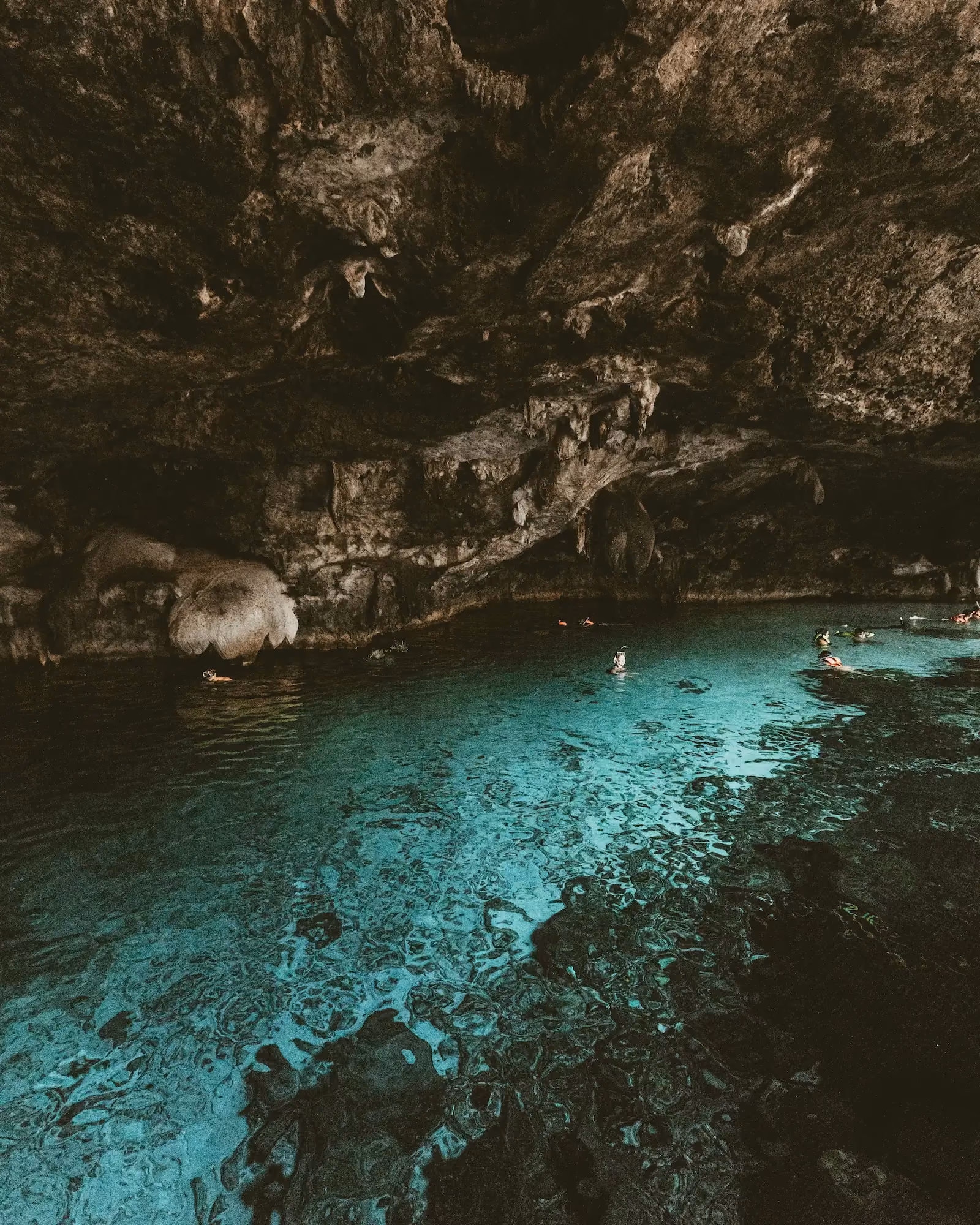 People snorkeling in clear turquoise water inside a large natural limestone cave with rugged rock walls and ceiling overhead.