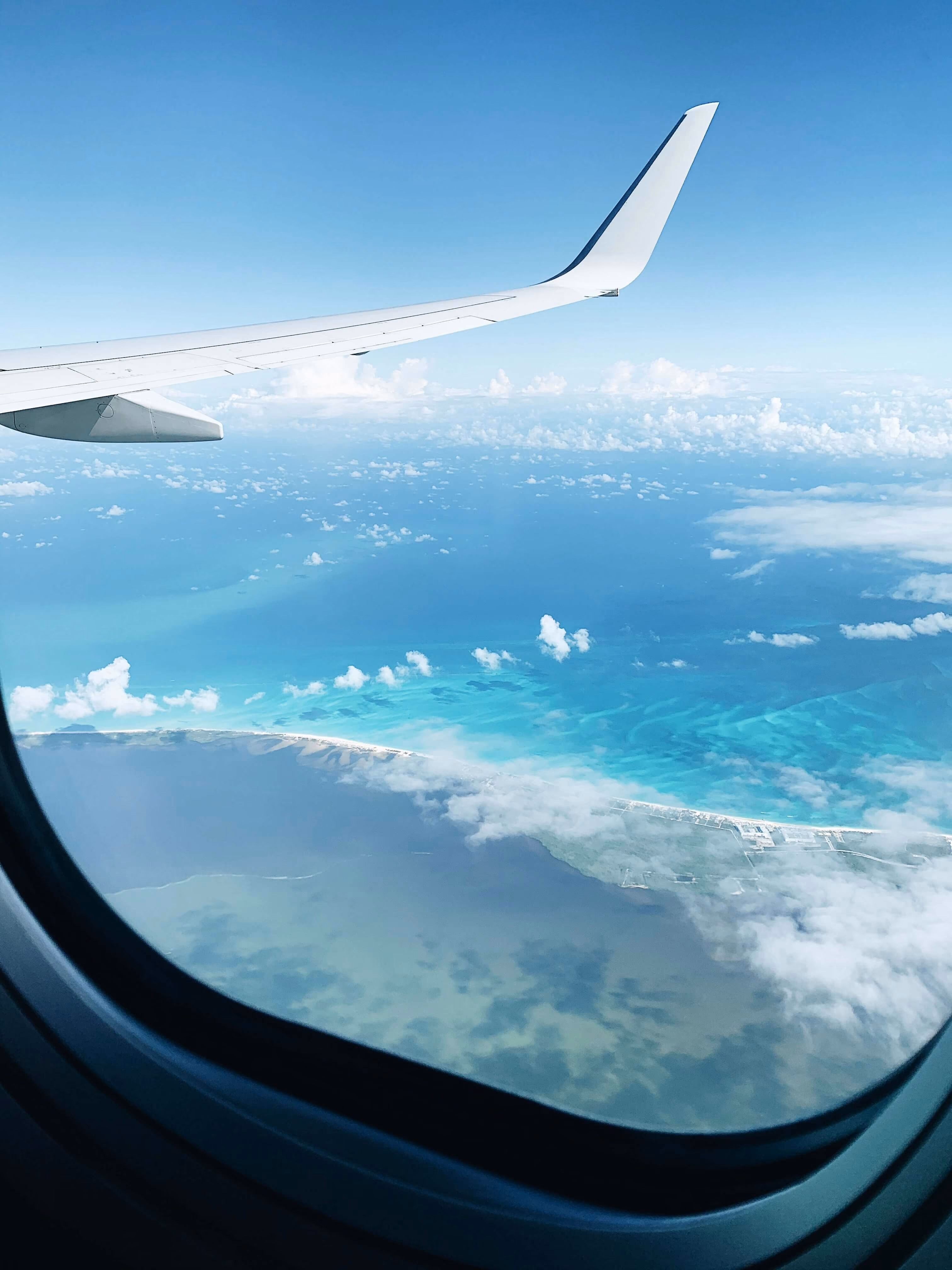 View from an airplane window showing the wing and a coastline with turquoise ocean waters, white sandy beaches, clouds, and clear blue sky.