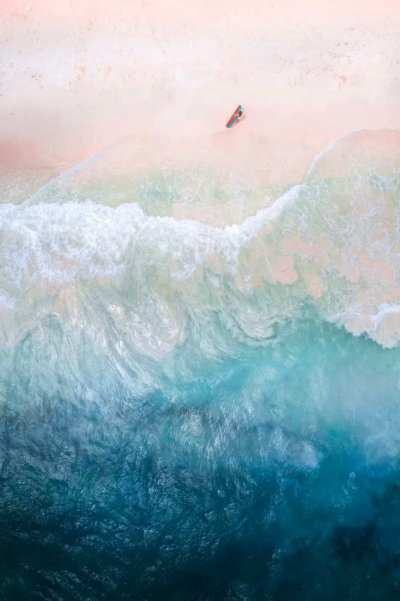 Aerial view of a person standing on a light sandy beach holding a red and teal surfboard near the foamy edge of turquoise ocean waves.