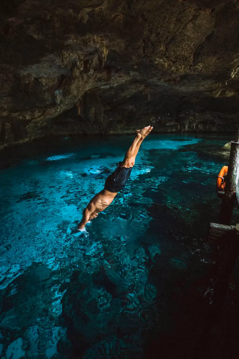 Man diving headfirst into clear blue water inside a rocky cave with stalactites on the ceiling.