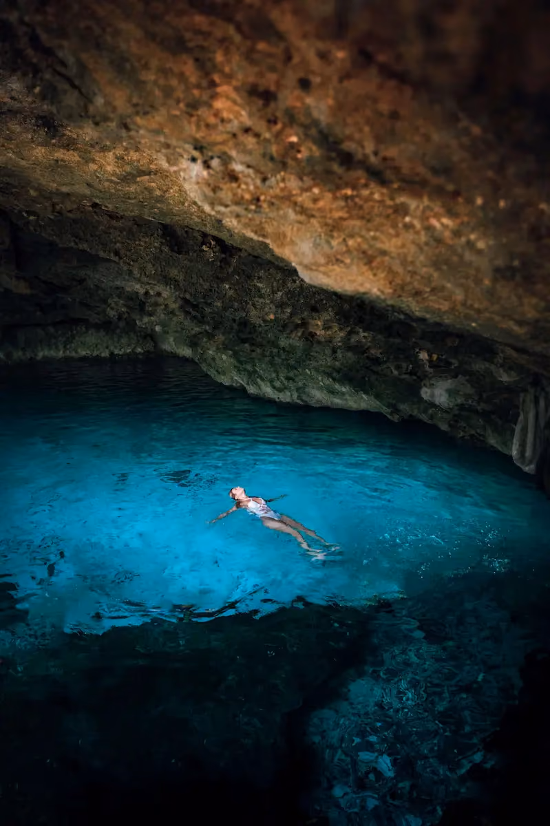 Person floating relaxed on their back in clear blue water inside a rocky cave with a textured ceiling.