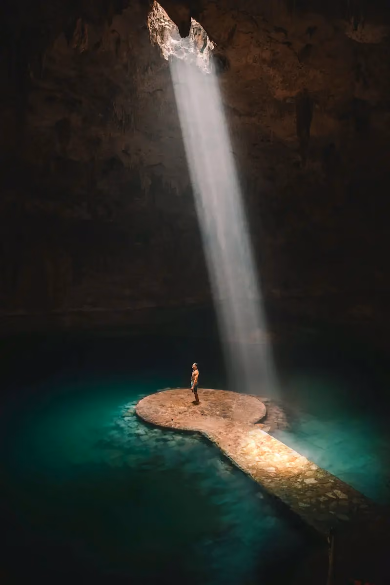 A person stands on a circular stone platform in the center of a turquoise underground pool inside a cave, illuminated by a strong beam of sunlight coming from an opening above.