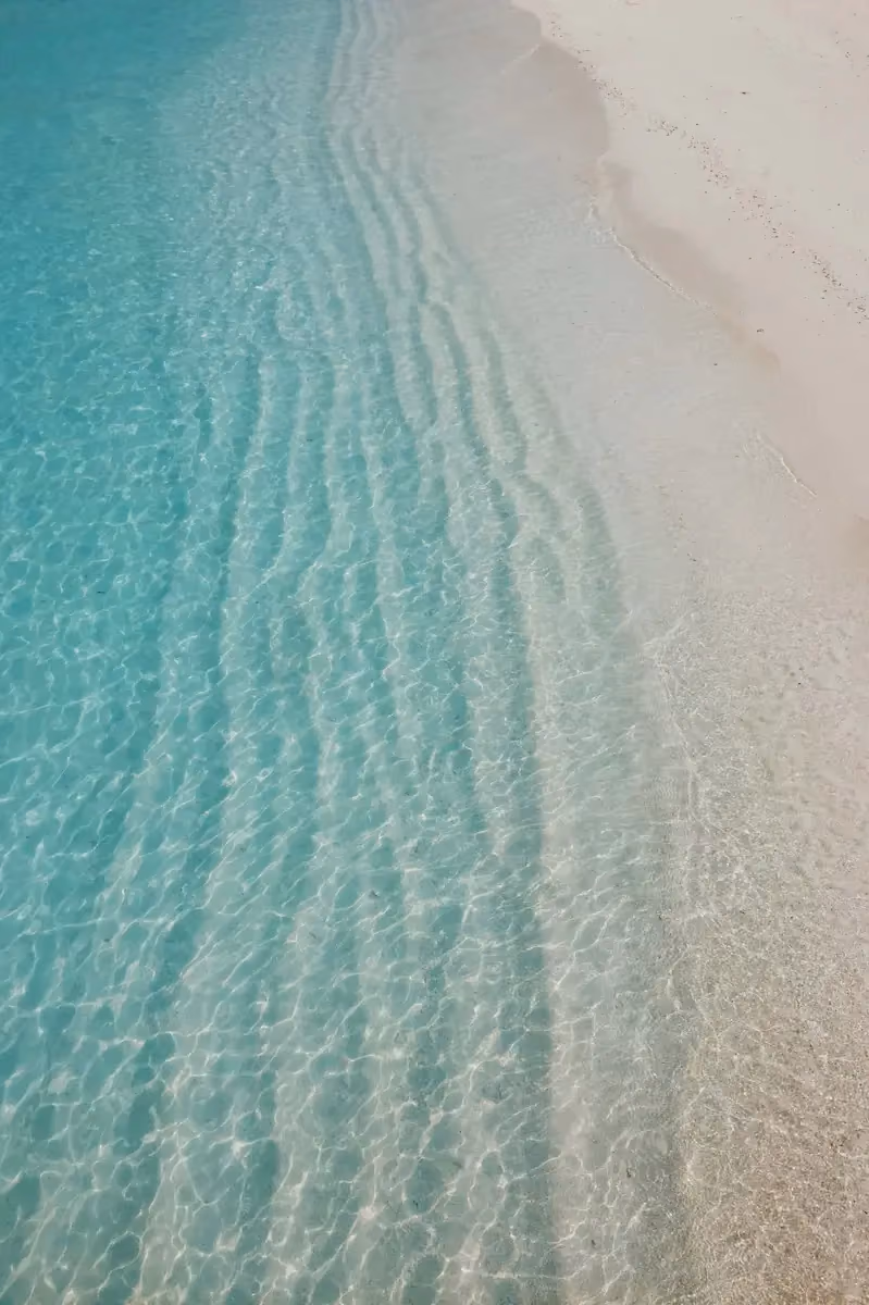 Clear turquoise ocean water gently washing onto a smooth sandy beach with ripple patterns visible beneath the shallow water.