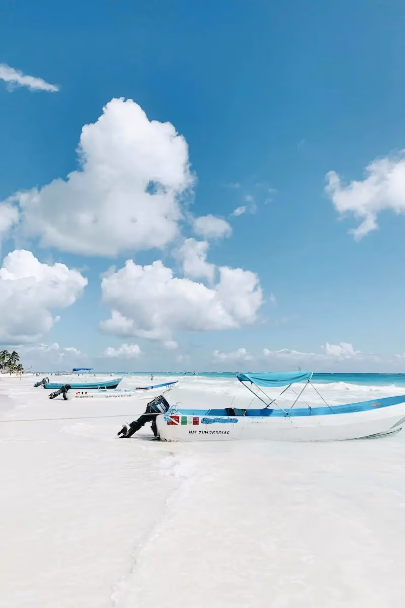 White sandy beach with several small fishing boats with blue trim and outboard motors resting at the shore under a bright blue sky with scattered fluffy clouds.