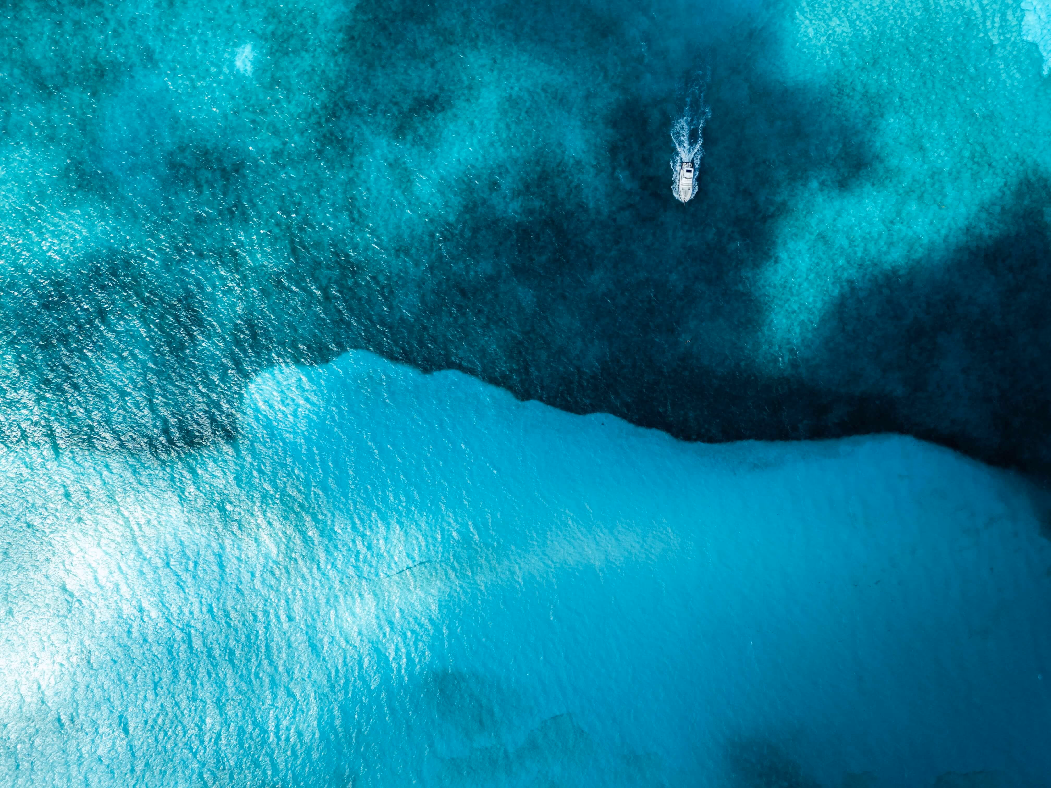 Aerial view of a small boat moving across clear turquoise ocean water with darker deep water patches and visible seabed texture.