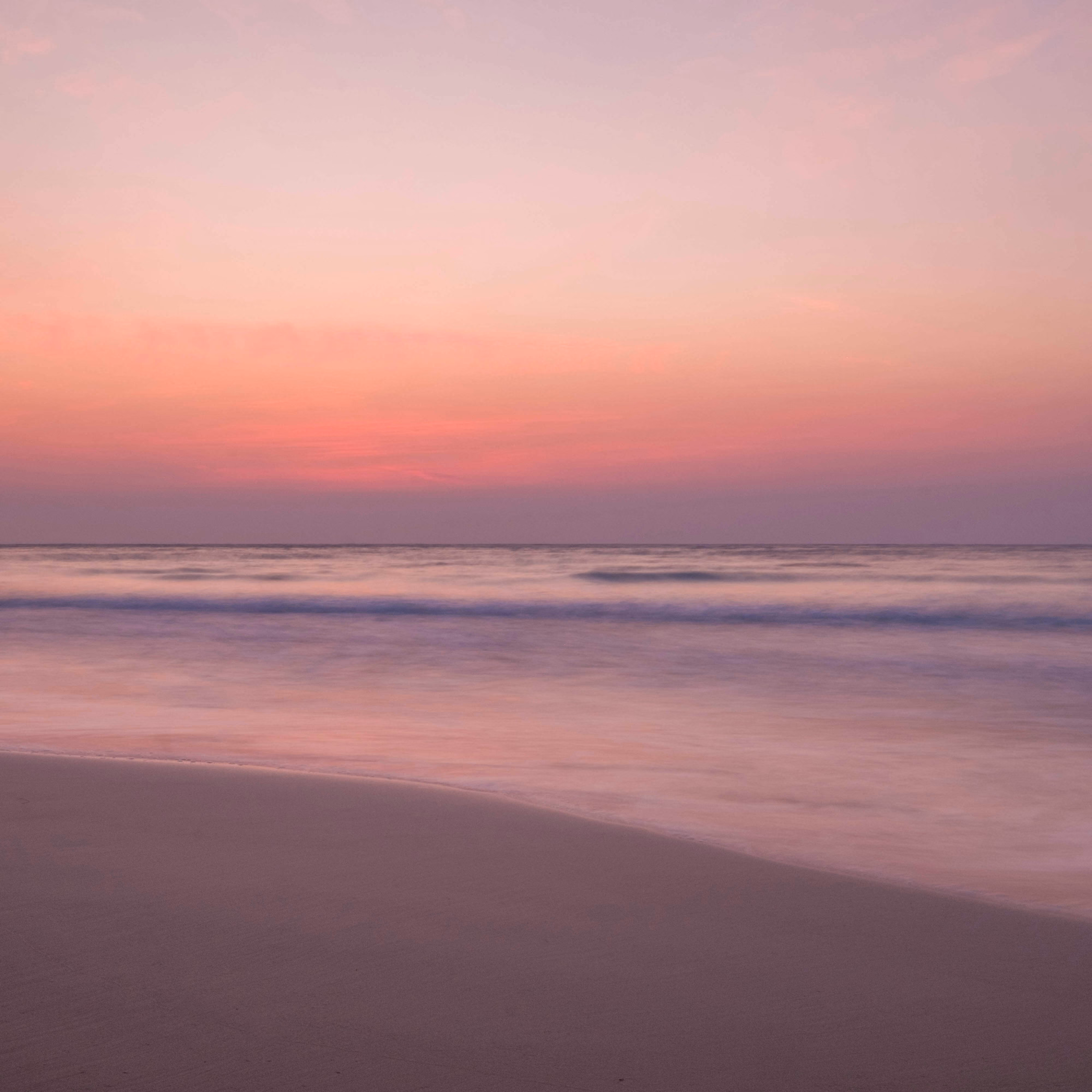 Peaceful beach scene at sunset with smooth gentle waves and a pink and purple sky over calm ocean water.