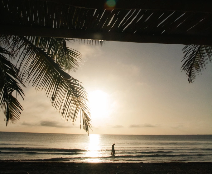 Silhouette of a person walking along a beach at sunset with calm waves reflecting the sun, framed by palm fronds and a shaded roof structure.