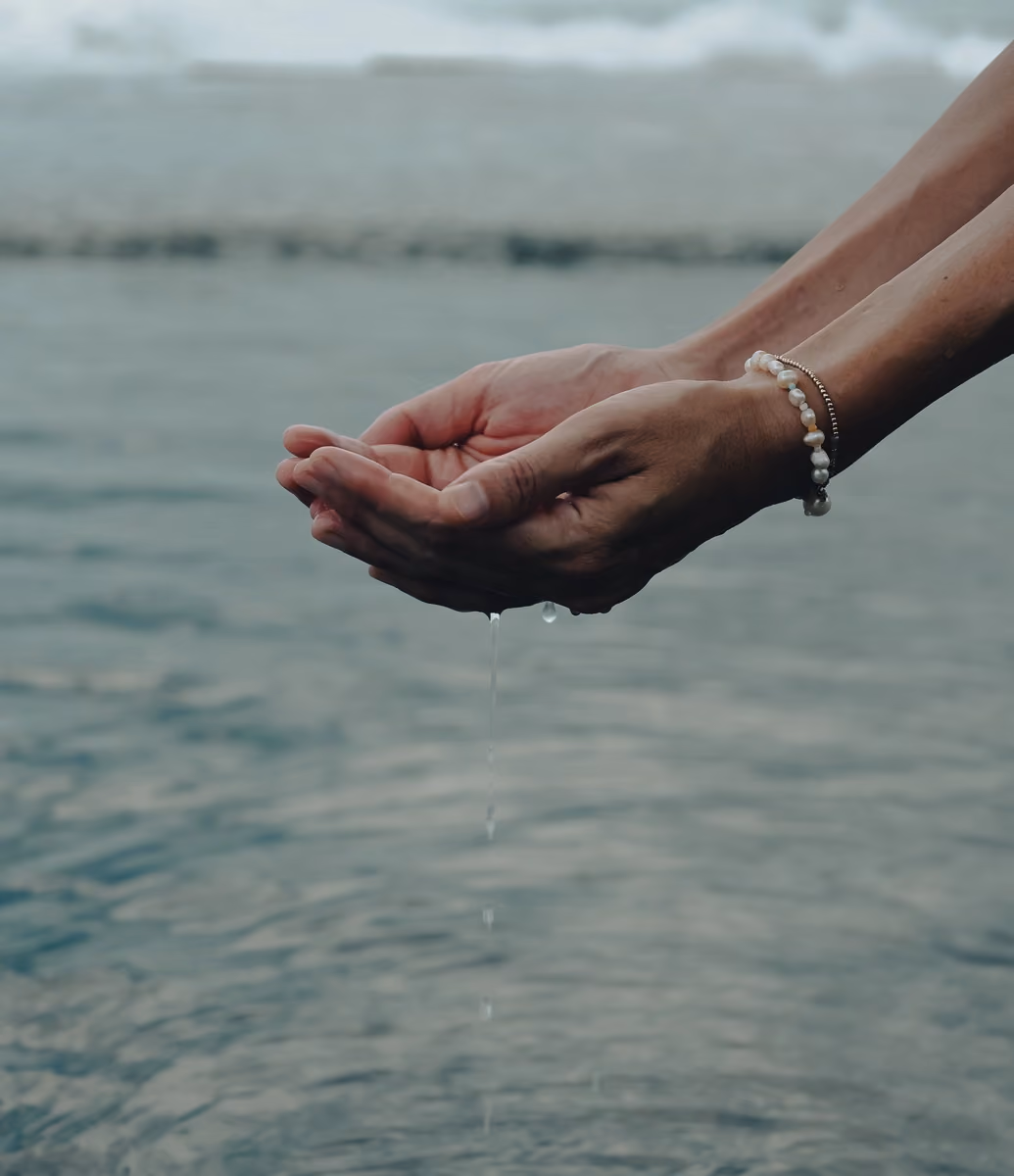 Two hands cupped together with water dripping from them over a body of water, one wrist adorned with a pearl bracelet and a thin gold chain bracelet.