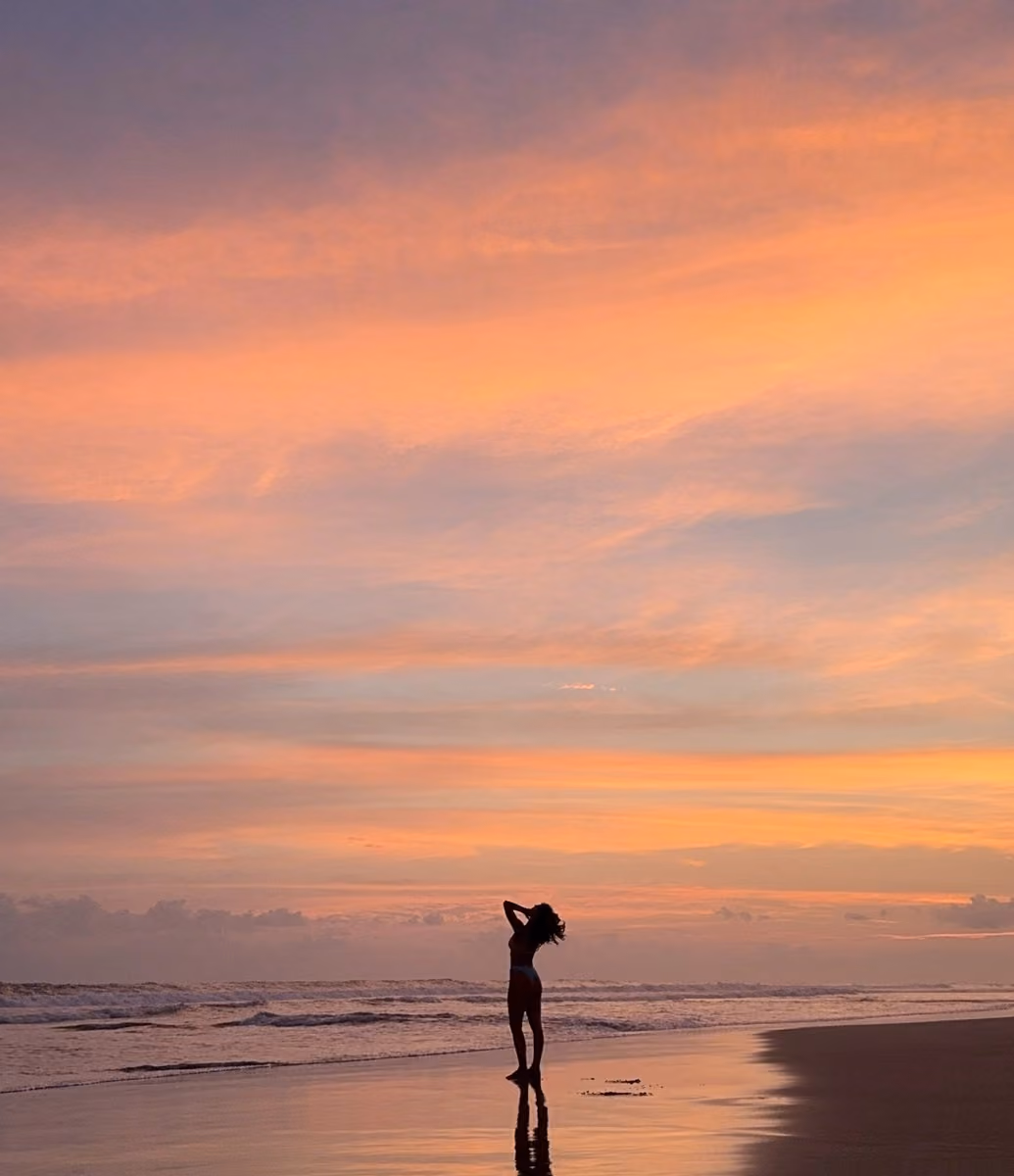 Silhouette of a woman standing on a wet beach at sunset with vivid orange, pink, and purple sky and gentle waves in the background.