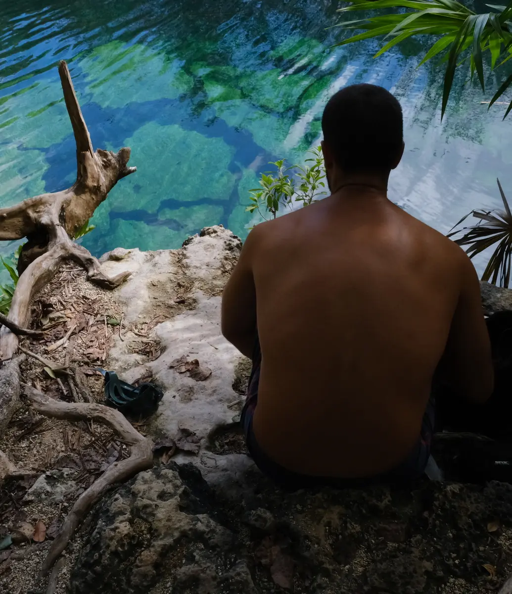 A shirtless person sitting on rocky ground near clear blue water with reflections, surrounded by leafy plants and tree branches.