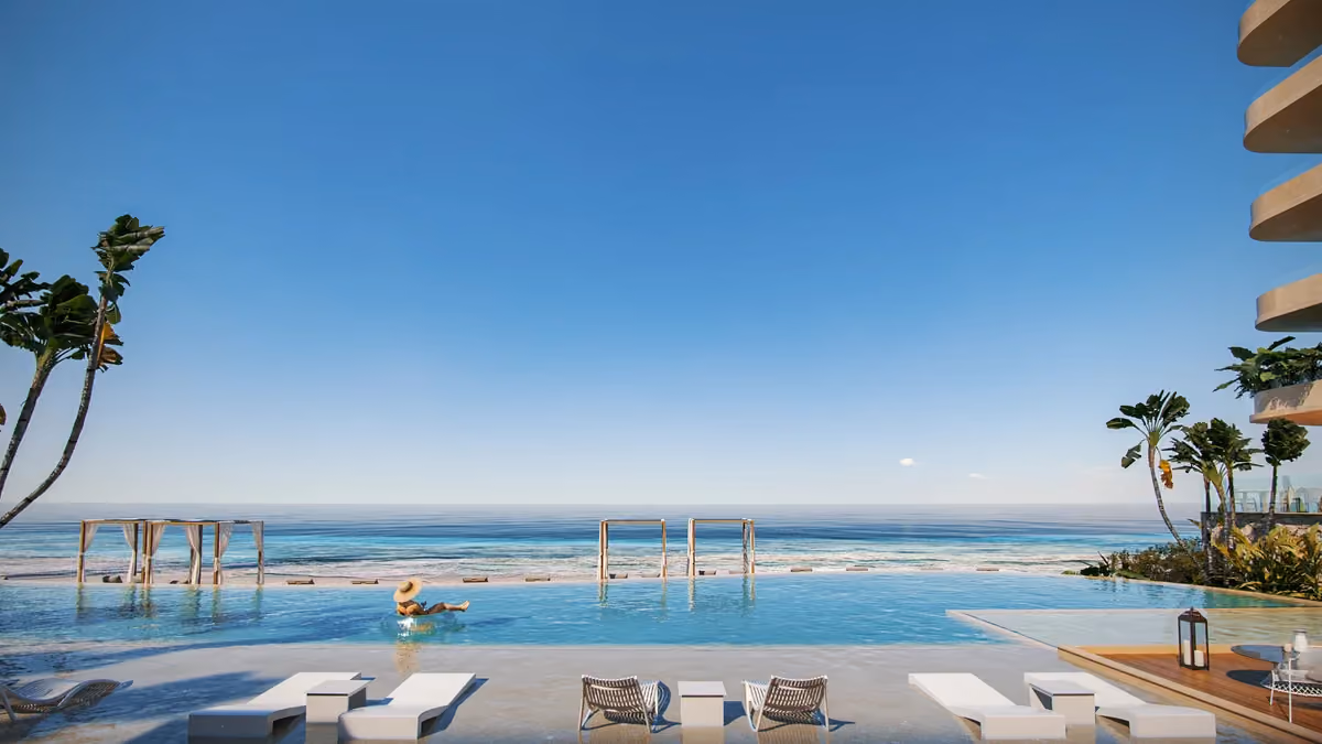 Infinity pool overlooking calm ocean under clear blue sky with lounge chairs and a person floating in the pool wearing a straw hat.