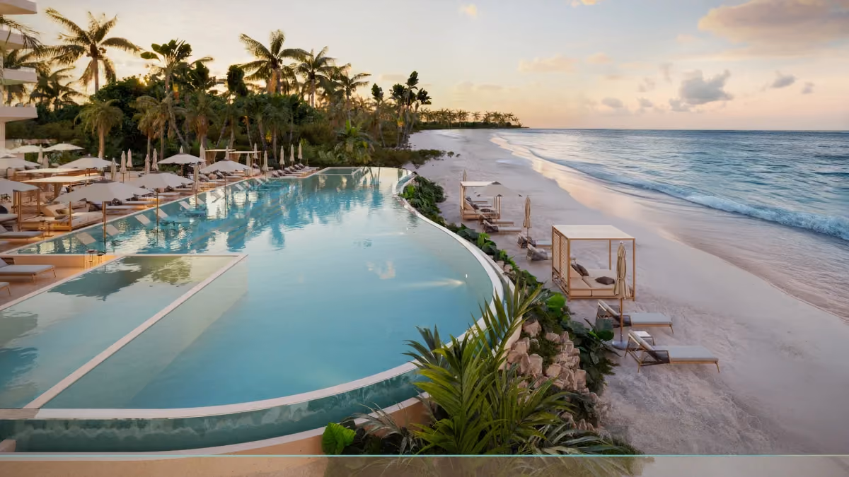 Infinity pool at a beach club with lounge chairs and umbrellas on the left side and cabana-style seating with sun loungers on the sandy beach to the right, overlooking a calm ocean under a partly cloudy sky at sunset.