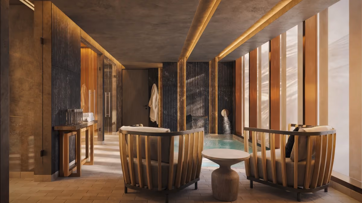 Modern spa room with a small indoor pool featuring water streams, two cushioned wooden lounge chairs, a round wooden side table, warm vertical wall panels, and soft ceiling lighting.