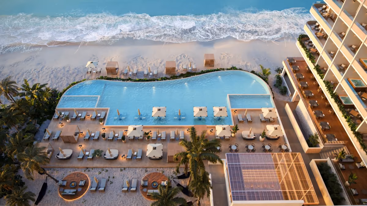 Aerial view of a beach resort with a large, irregularly shaped swimming pool surrounded by lounge chairs, umbrellas, and cabanas, adjacent to a sandy beach with ocean waves.
