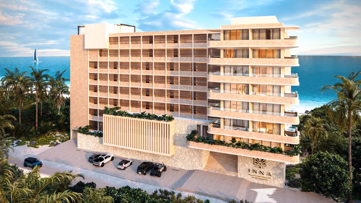 Modern multi-story beachfront building with balconies, surrounded by palm trees and parked cars, overlooking a calm ocean with a sailboat in the distance under a partly cloudy sky.