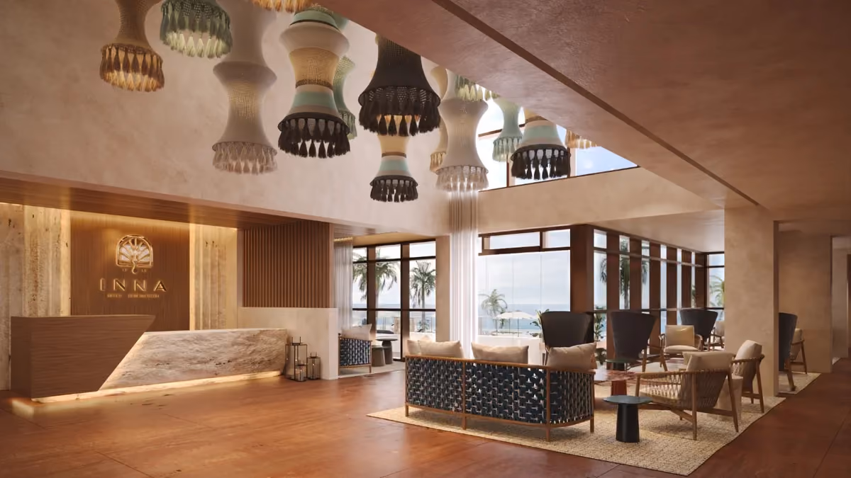 Modern hotel lobby with wooden floor and reception desk illuminated by soft lighting, featuring large windows with ocean views, woven pendant lamps hanging from the ceiling, and seating area with patterned sofas and wooden chairs on rugs.