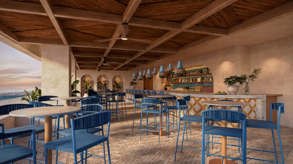 Modern beach club bar with blue chairs and round wooden tables under a wooden beam ceiling, overlooking the ocean at sunset.