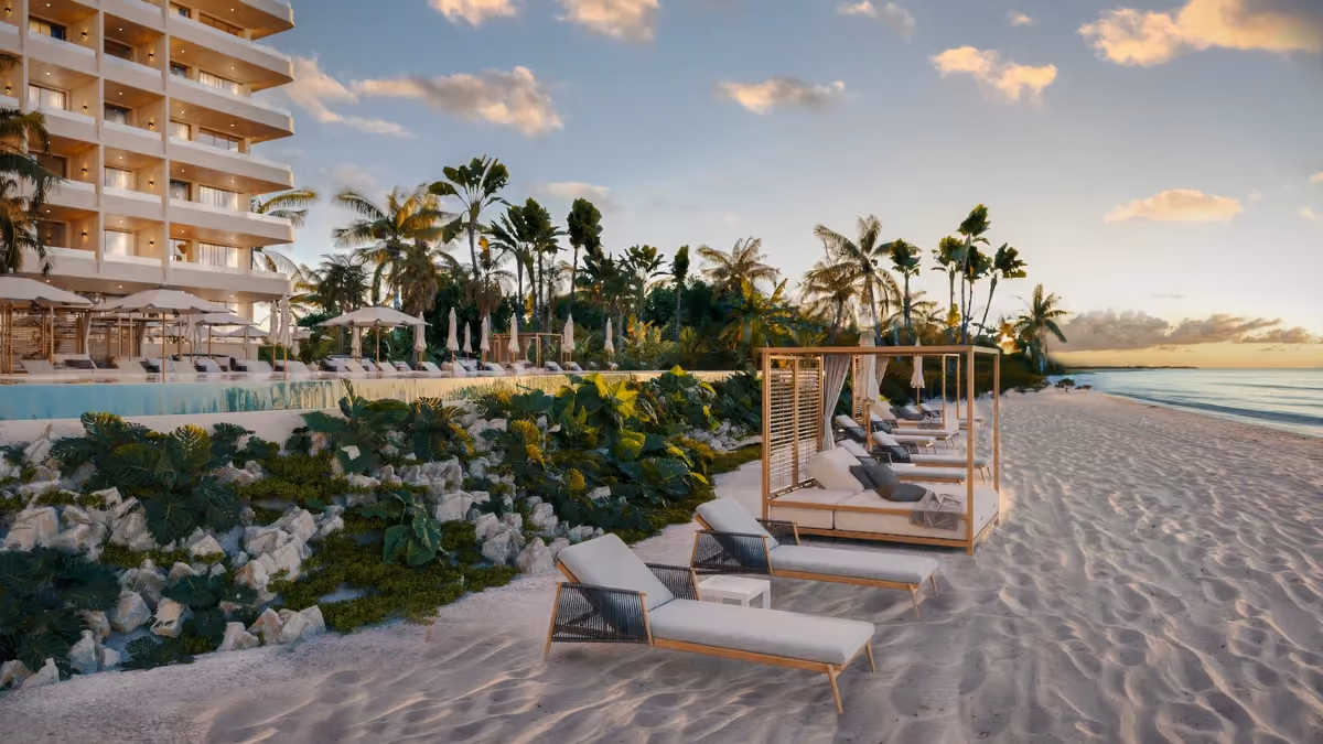 Beachside view of a luxury resort at sunset with lounge chairs and cabanas on the sandy shore, tropical plants, palm trees, a pool, and a multi-story building with balconies on the left.