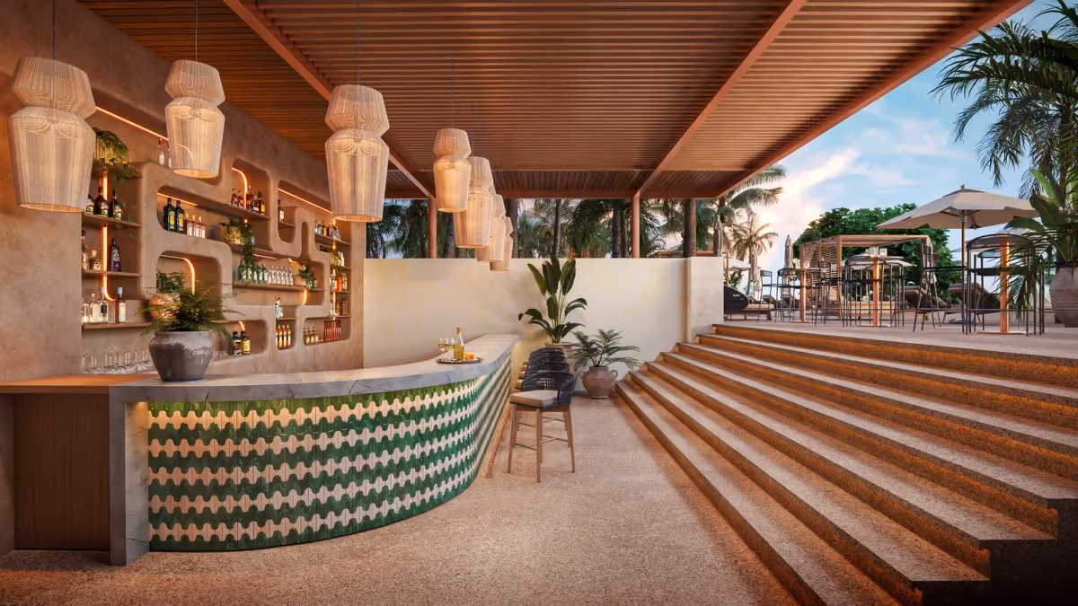 Modern outdoor snack bar with a curved marble counter featuring green patterned tiles, hanging woven lamps, and shelves with bottles and plants, next to a stairway leading up to a patio area with tables, umbrellas, and lush palm trees.