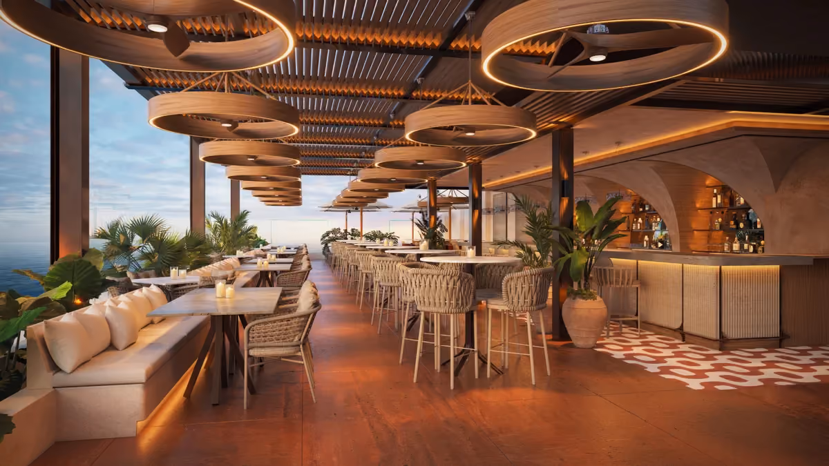 Elegant rooftop bar and dining area with round wooden pendant lights, wicker chairs, cushioned bench seating along the left side, tables with candles, large plants, a bar with shelves of bottles, and a view of the ocean under a partly cloudy sky.