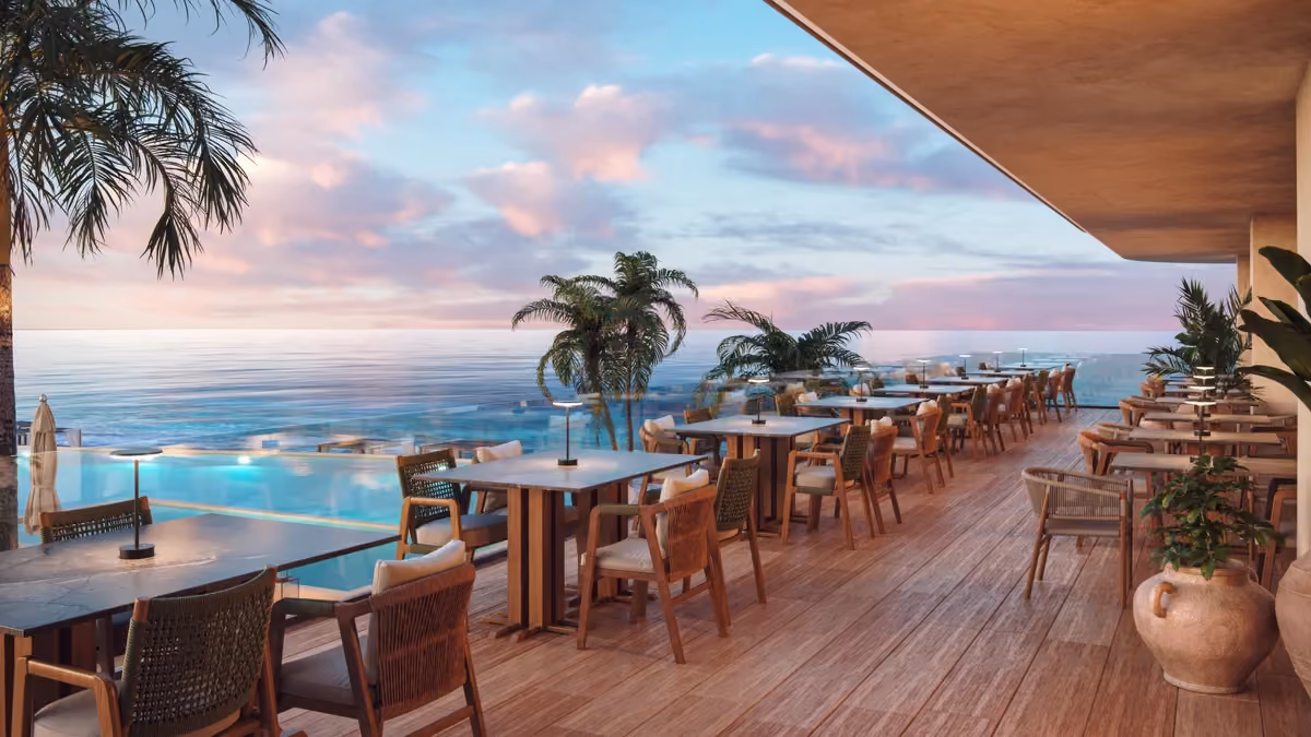 Outdoor restaurant terrace with wooden tables and cushioned chairs overlooking an infinity pool and ocean under a partly cloudy sky at sunset.