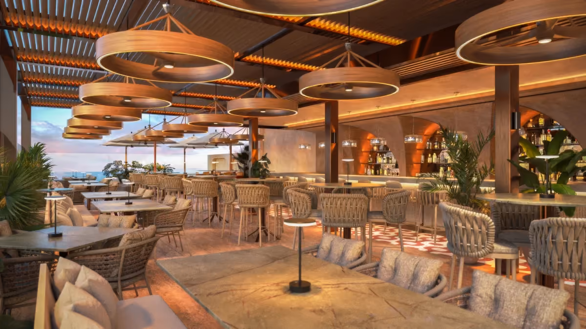 Modern rooftop restaurant with woven chairs, cushioned seating, marble tables, large circular wooden pendant lights, plants, and a bar with shelves of bottles in the background.
