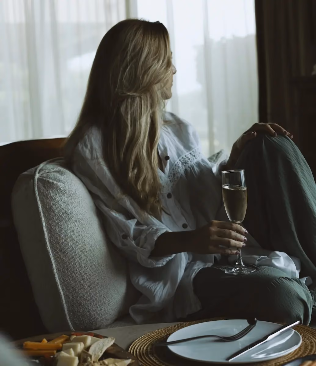 Woman with long blonde hair sitting on a couch near a window with sheer curtains, holding a glass of champagne and looking to the side, with a plate and cutlery on a table and a cheese platter in the foreground.