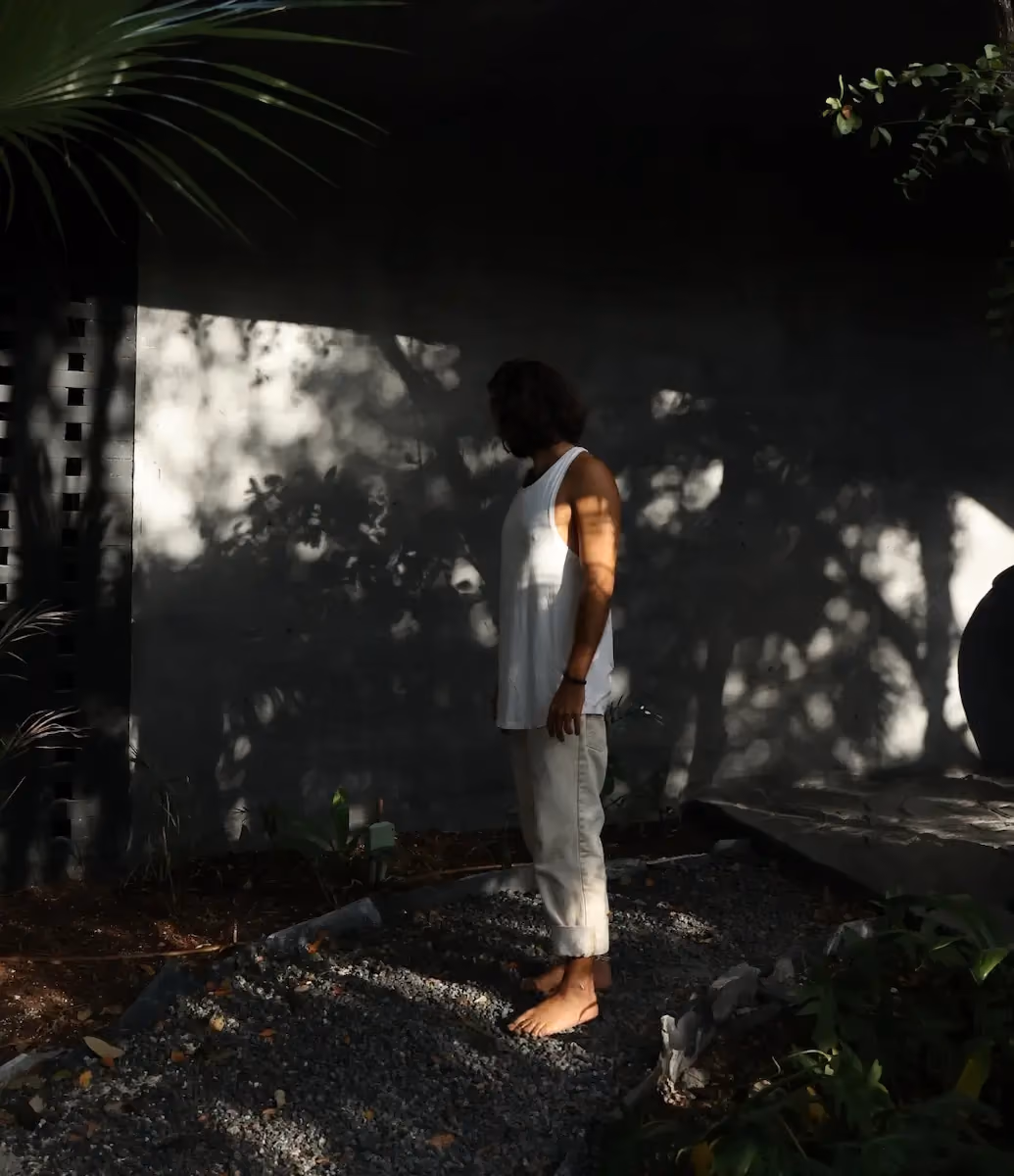 A barefoot person with shoulder-length hair wearing a white sleeveless top and light-colored rolled-up pants stands on gravel next to a garden bed, with shadows of foliage cast on a wall behind them.
