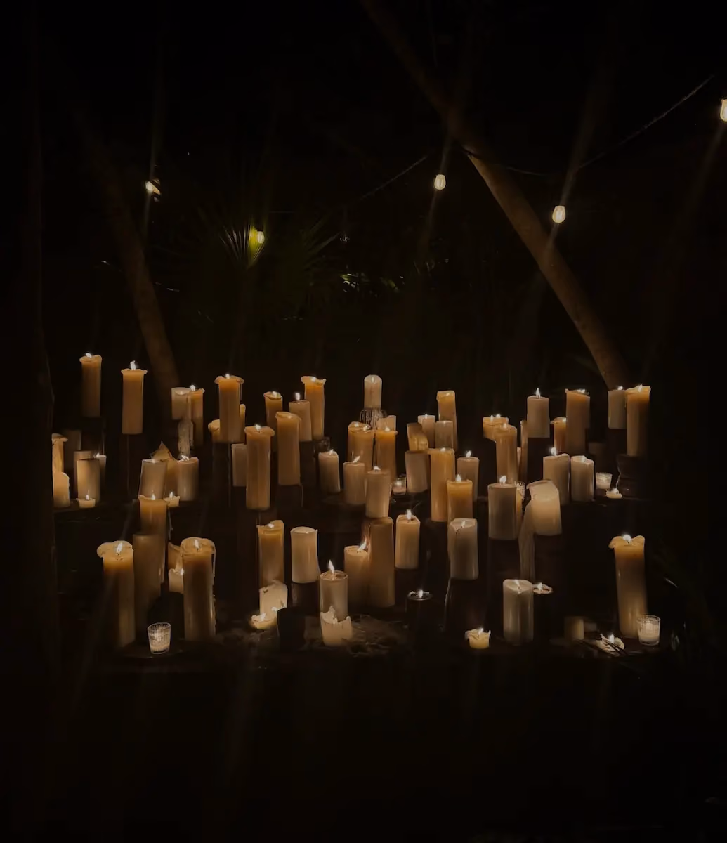 Numerous lit candles of various sizes arranged outdoors on a wooden surface at night, softly illuminating the surrounding area with warm light among trees and string lights.