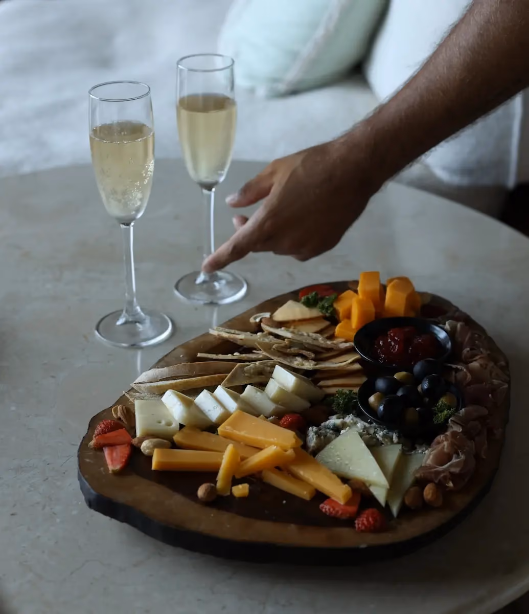 A hand reaching for a glass of sparkling wine on a table next to a wooden board with assorted cheeses, crackers, olives, fruits, and nuts.