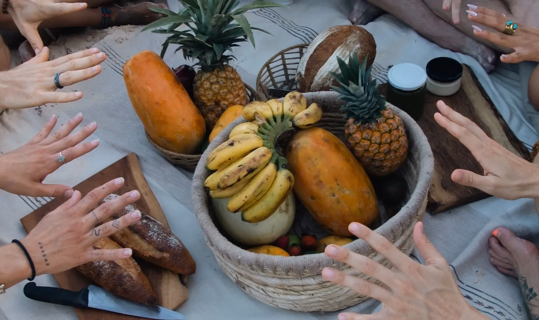 A top-down view of a picnic setup with a woven basket containing bananas, pineapples, papayas, and other fruits, surrounded by hands reaching toward the food, with bread on a wooden board and jars on a light-colored cloth.