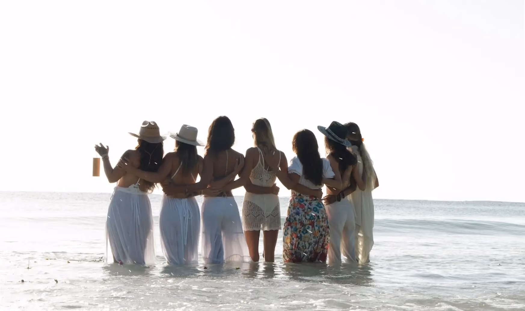 Six women standing in shallow ocean water with their backs to the camera, arms around each other, wearing summery dresses and hats, facing the horizon at sunset.