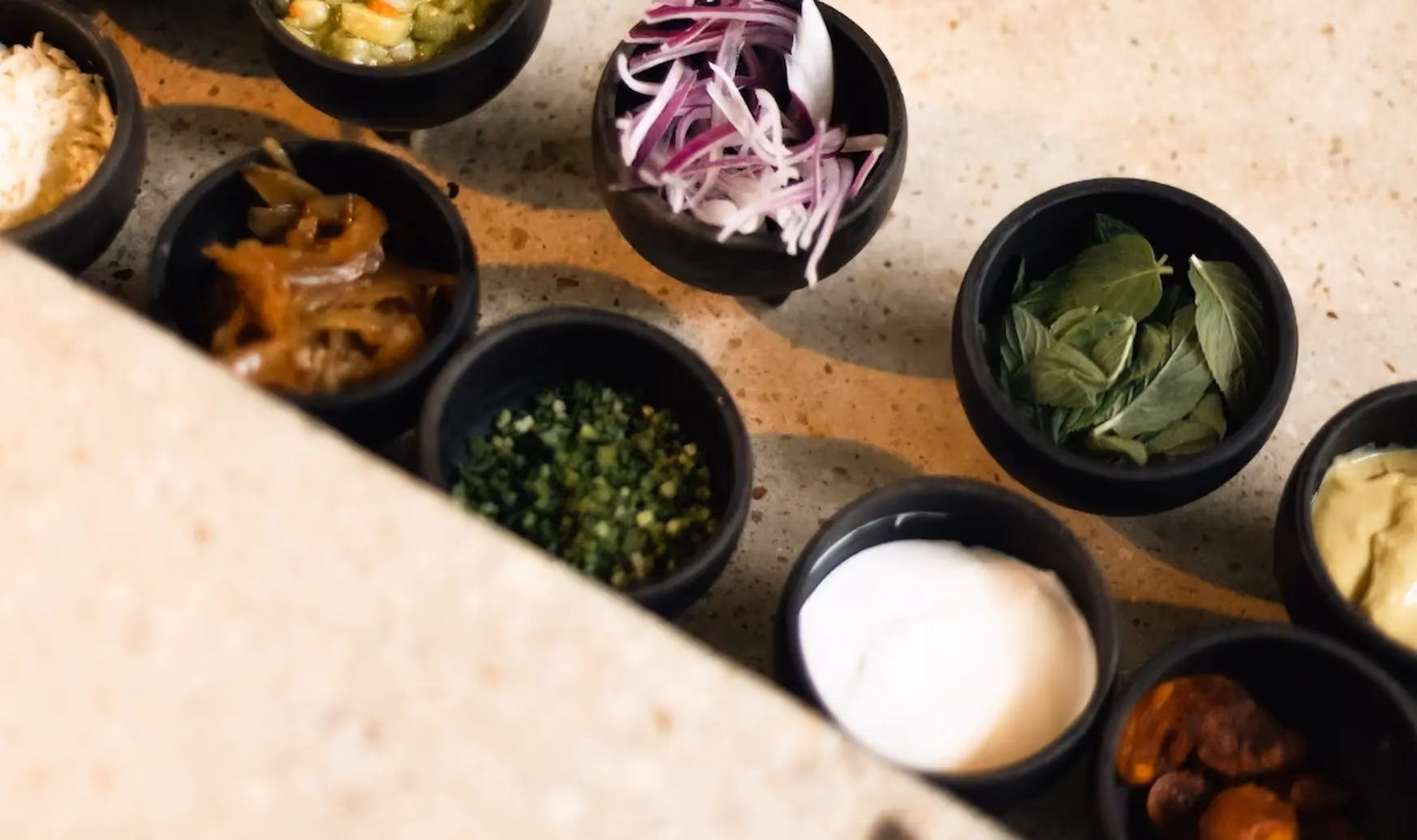 Small black bowls with various ingredients including sliced red onions, fresh green herbs, white sauce, chopped green herbs, cooked onions, and other sauces on a beige countertop.