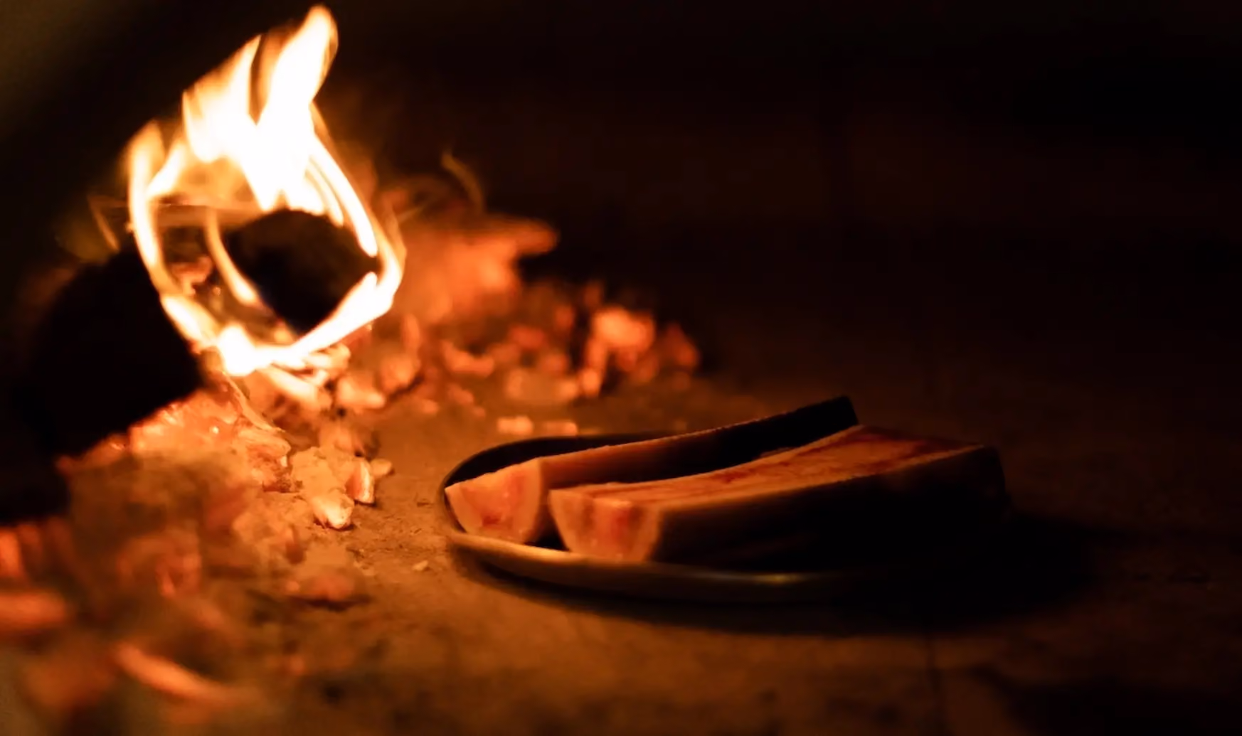 Two raw bone marrow halves on a plate near glowing firewood and embers inside a dark fireplace.