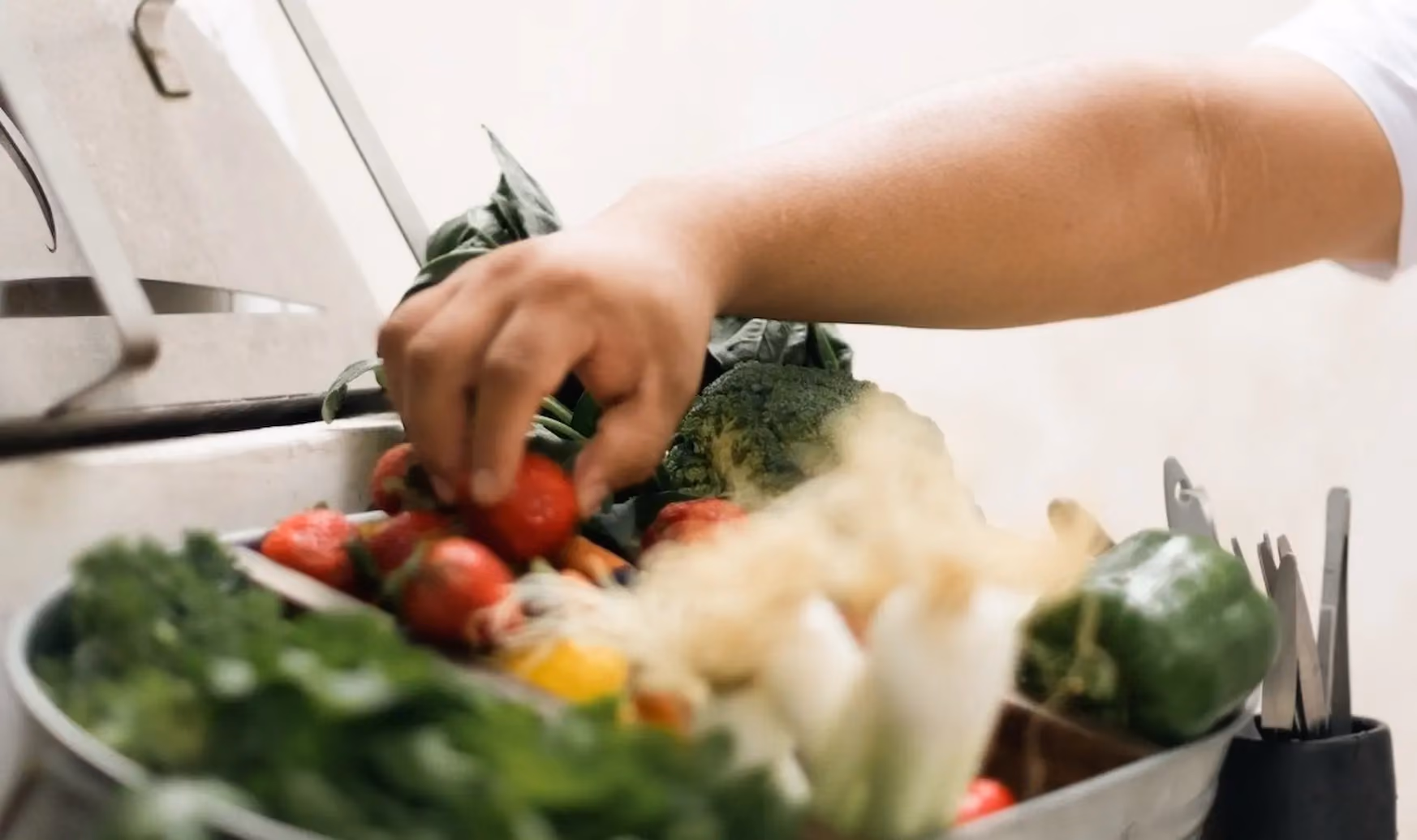 Person's hand reaching to pick a strawberry from a bowl filled with various fresh vegetables and fruits including broccoli, leafy greens, green bell pepper, and carrots.