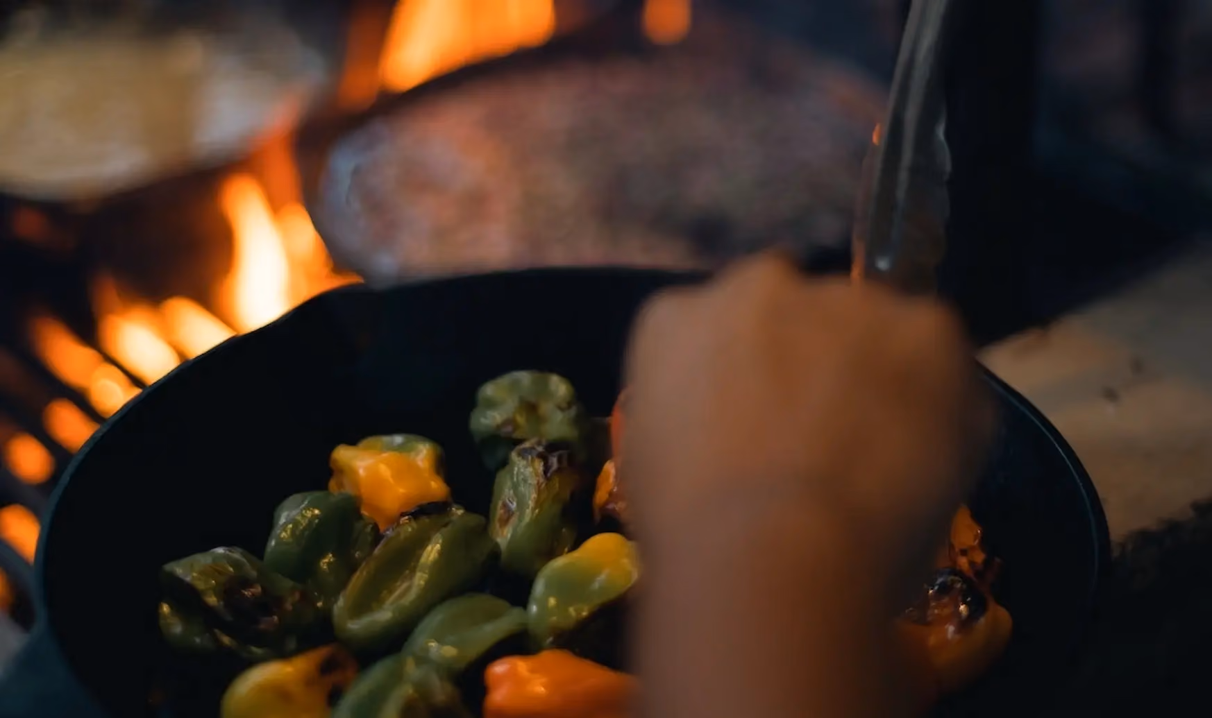 Hand holding tongs roasting green and orange peppers over a flame in a black pan.