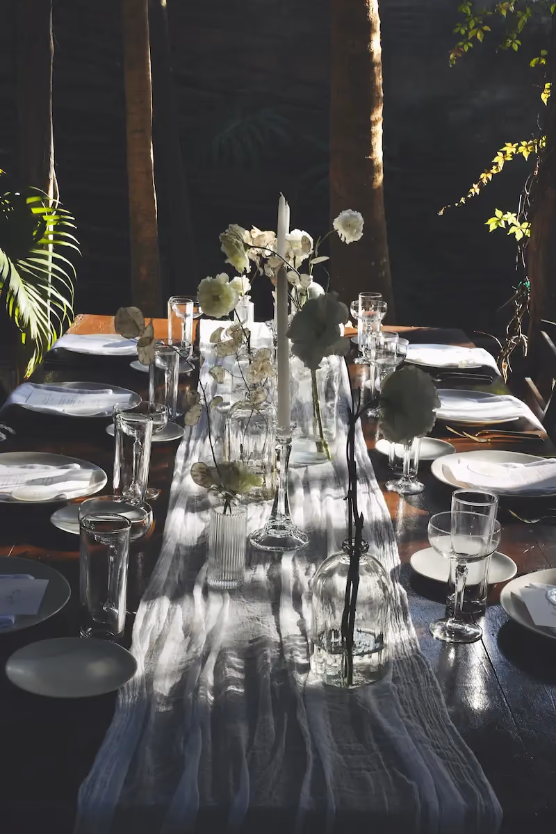 Elegant dining table with a white textured runner, clear glass vases holding white flowers, glass candleholder with a white candle, and neatly arranged white plates and glassware, set in a softly lit room with trees and greenery in the background.