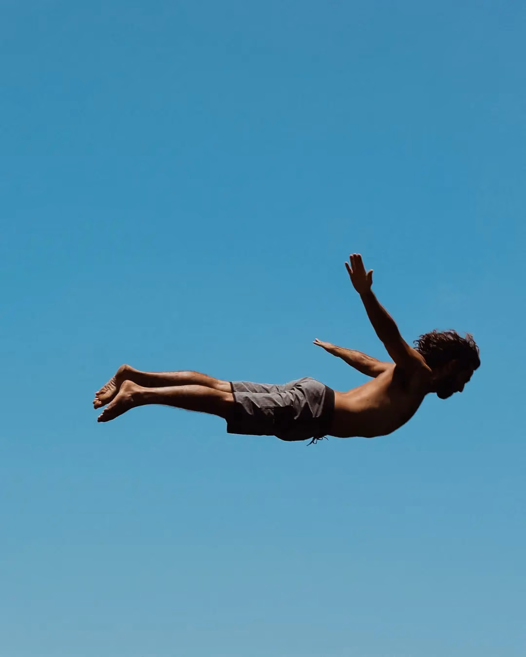 A shirtless man in gray shorts is captured mid-air against a clear blue sky, appearing to be diving or falling with arms extended backward.