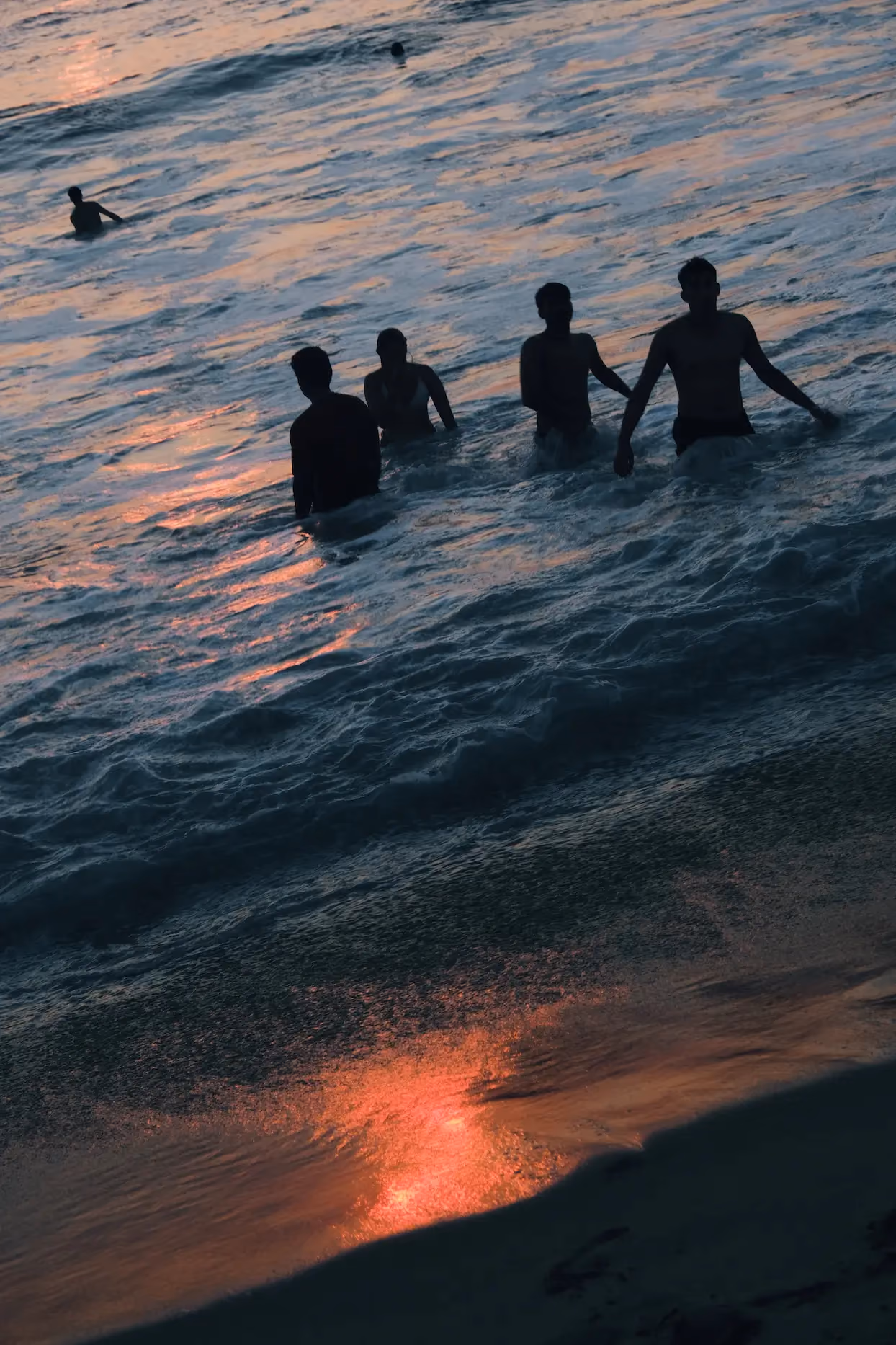 Silhouettes of five people standing waist-deep in ocean waves at sunset, with orange sunlight reflecting on water and wet sand.