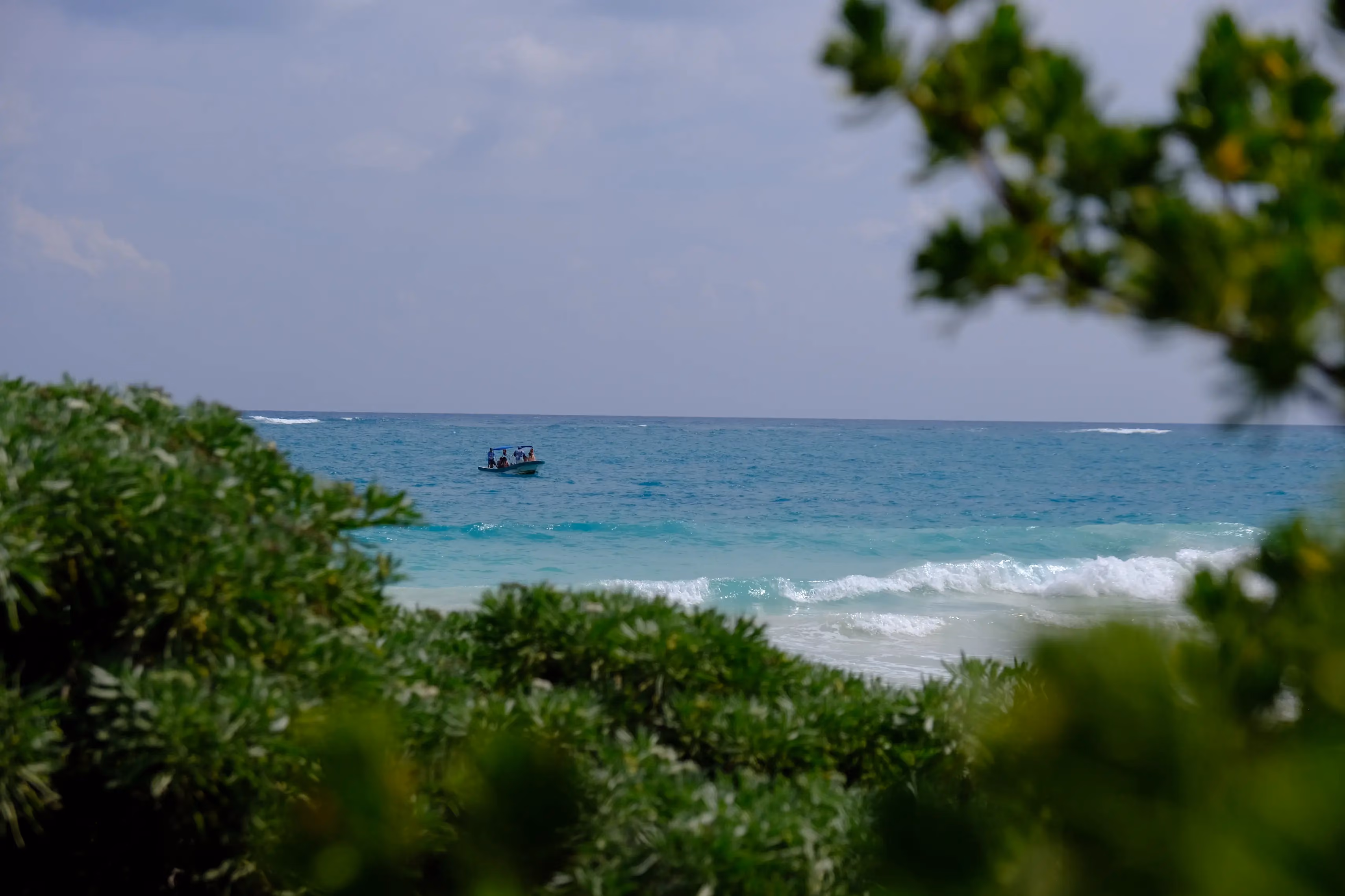 Small boat with people on turquoise ocean water seen through green coastal vegetation under a partly cloudy sky.
