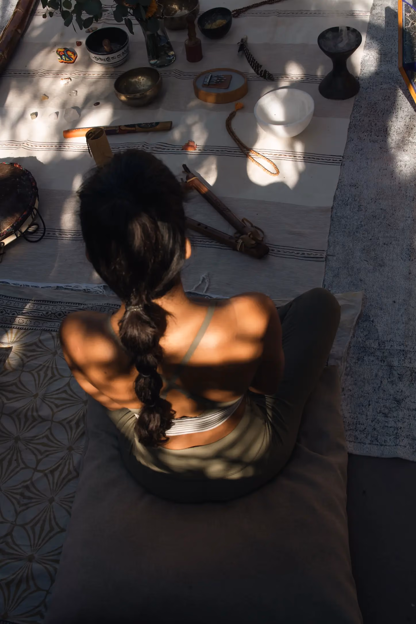 A woman with a braided ponytail sits cross-legged on a cushion, seen from behind, surrounded by various musical instruments and ritual objects on a striped cloth under soft shadow patterns.