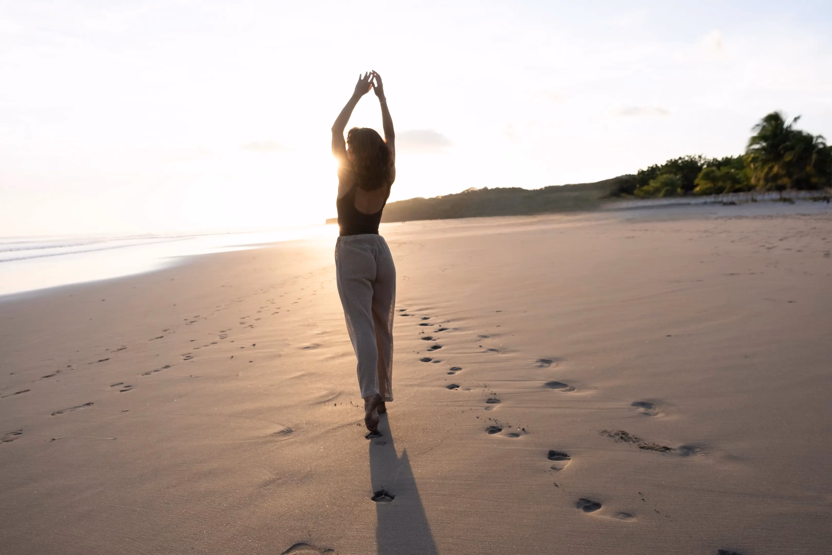 A woman with curly hair wearing a black top and light pants walks barefoot on a sandy beach at sunset, with her arms raised above her head and footprints trailing behind her.