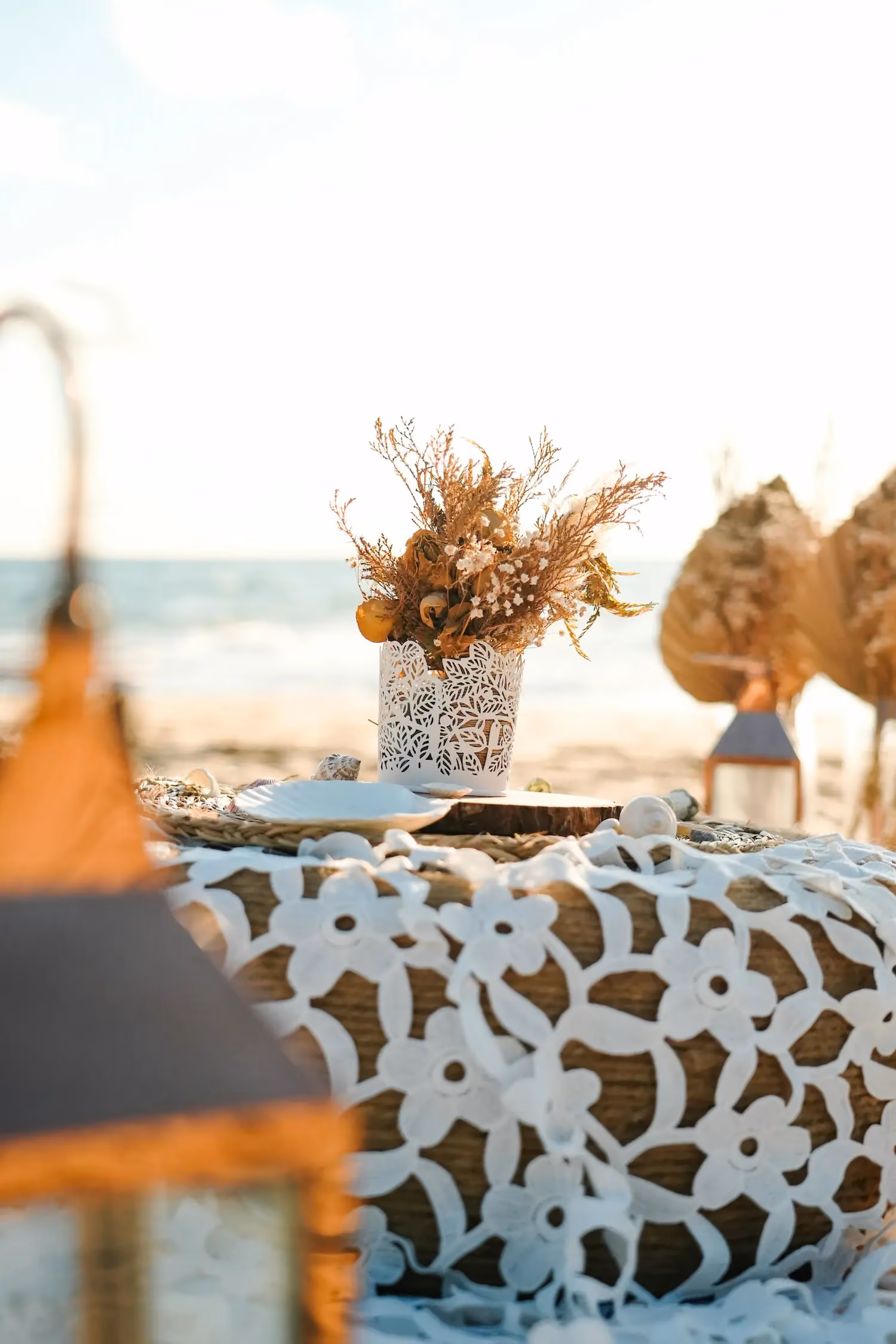 A white lace tablecloth with floral patterns covers a table set on a beach, featuring a white vase with dried flowers and surrounding seashells, with the sea visible in the background.