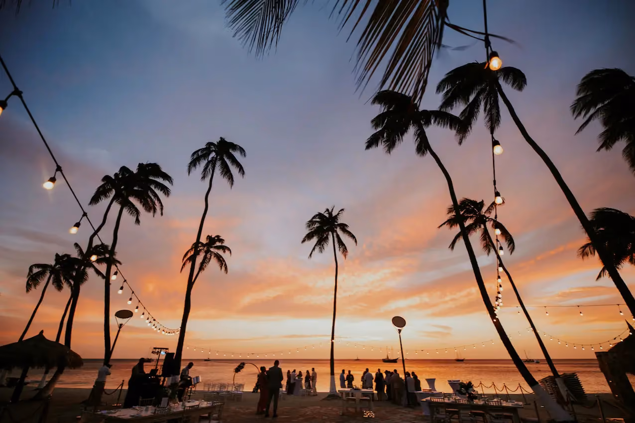 Silhouettes of people gathered on a beach under tall palm trees with string lights during a colorful sunset over the calm ocean.
