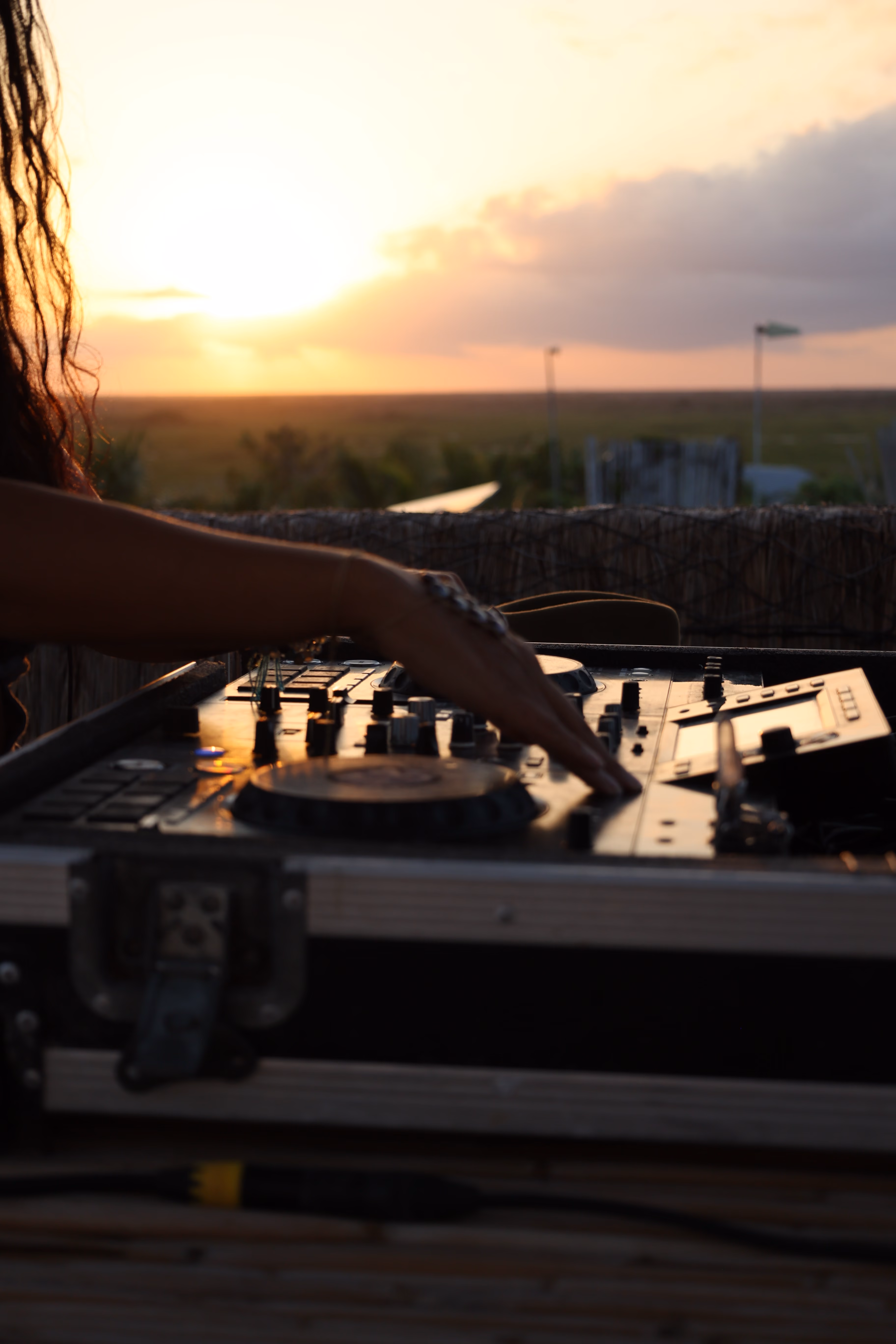 A person operating a DJ mixer and controller with one hand during sunset, with an outdoor landscape and partly cloudy sky in the background.