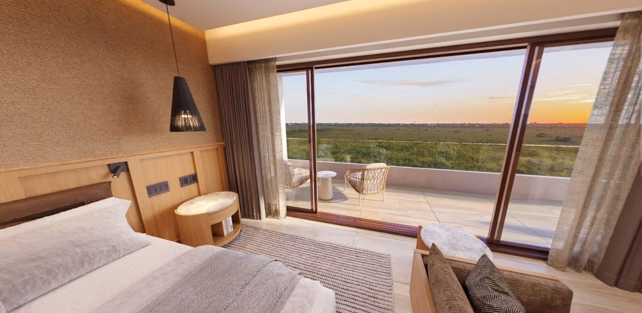 Modern bedroom with a large bed, cozy pillows, wooden side tables, a hanging black lamp, and floor-to-ceiling glass doors opening to a balcony with two wicker chairs and a small table overlooking a green landscape at sunset.