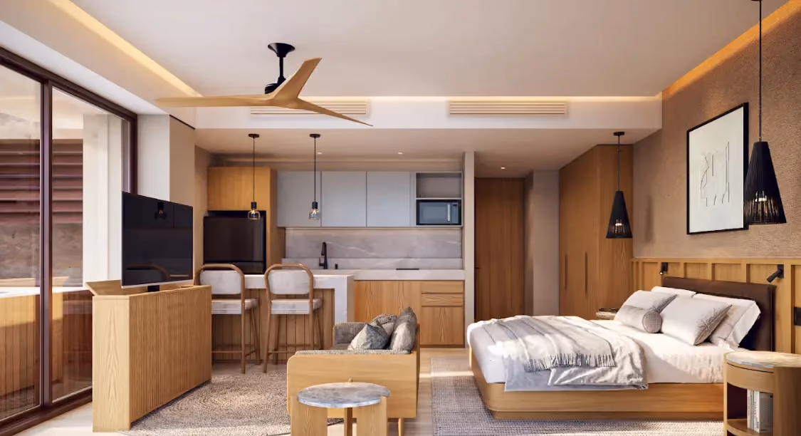 Modern studio apartment with wood accents featuring a bed with white bedding, nightstands, pendant lights, a small sofa, round wooden table, and a kitchenette with bar stools, cabinets, and appliances, all illuminated by natural light from large sliding glass doors.