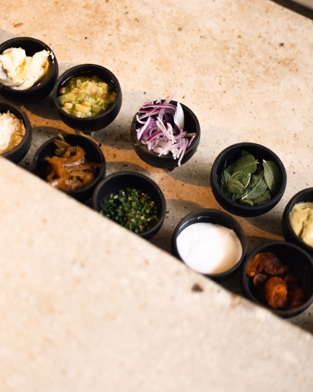 Small black bowls arranged on a light textured surface containing various condiments and ingredients, including cream, green salsa, sliced red onions, fresh green leaves, chopped herbs, caramelized onions, shredded cheese, white sauce, guacamole, and dried fruit pieces.
