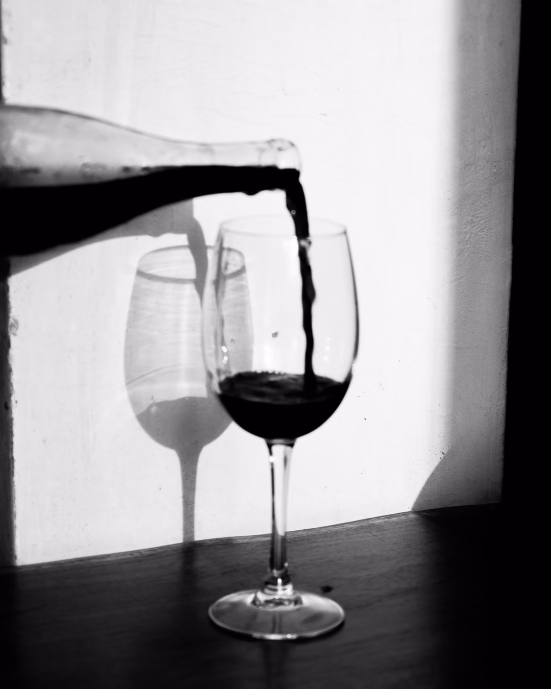 Black and white photo of red wine being poured from a bottle into a wine glass on a wooden surface, casting shadows on a white wall.